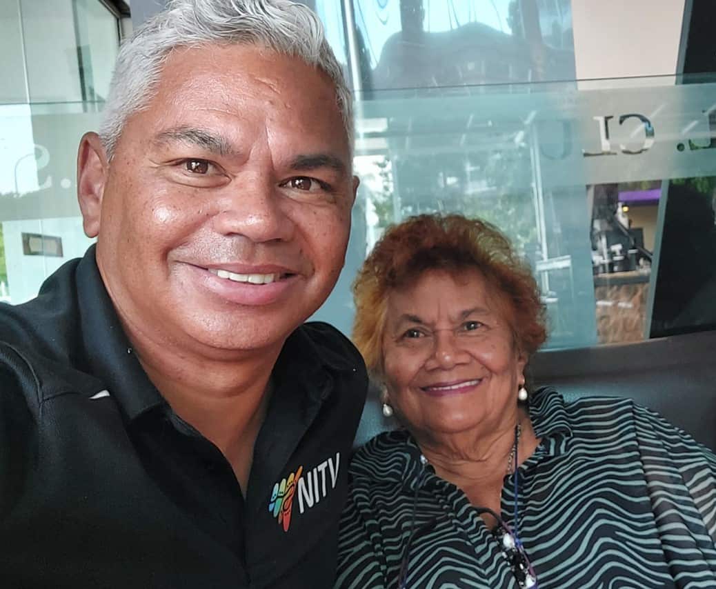 A man with grey hair smiles as he stands next to an elderly woman.