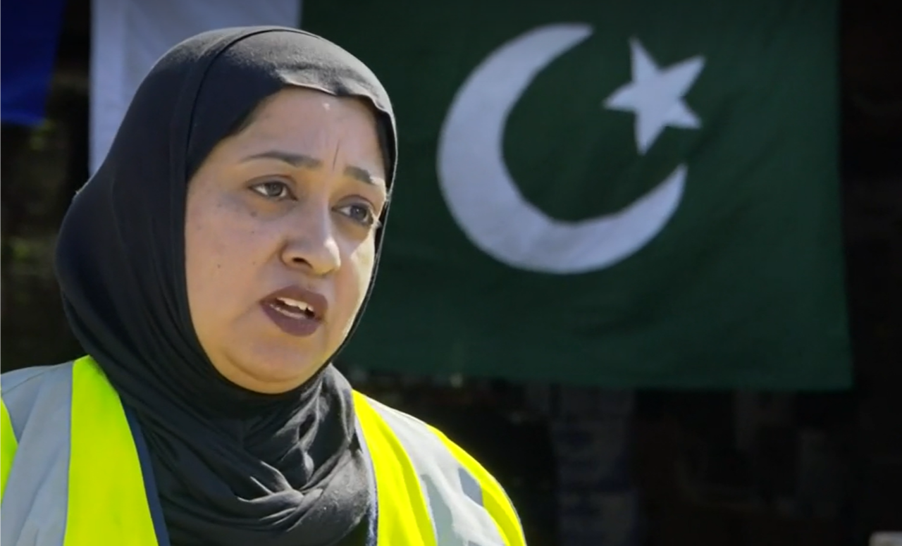 Woman in hijab and hi-vis vest stands in front of Pakistan flag. 