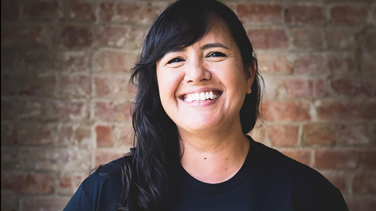 A woman with black hair, wearing a black t-shirt, standing in front of a brick wall. She is smiling.
