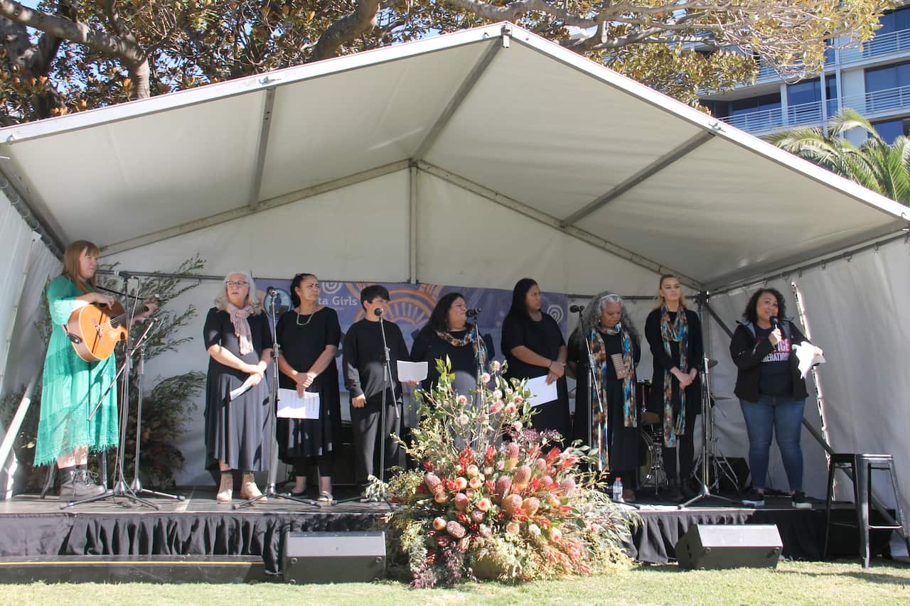 Nine women are standing on a stage under a white tent as part of a Reconciliation Week event. 
