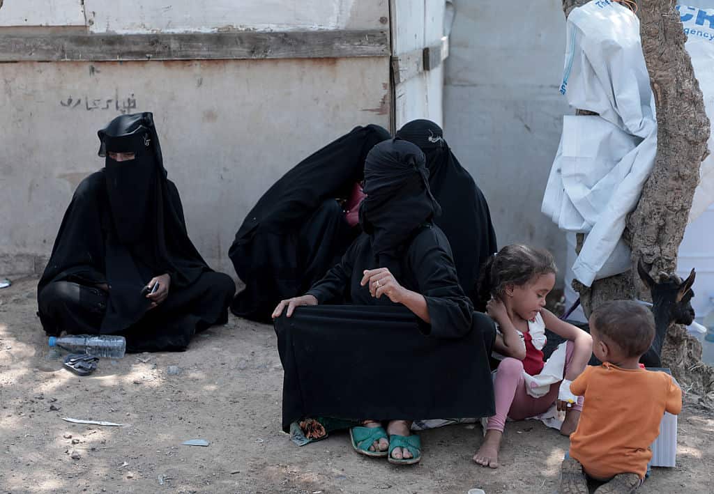 Women and children sit on the dirt outside temporary shelters. 