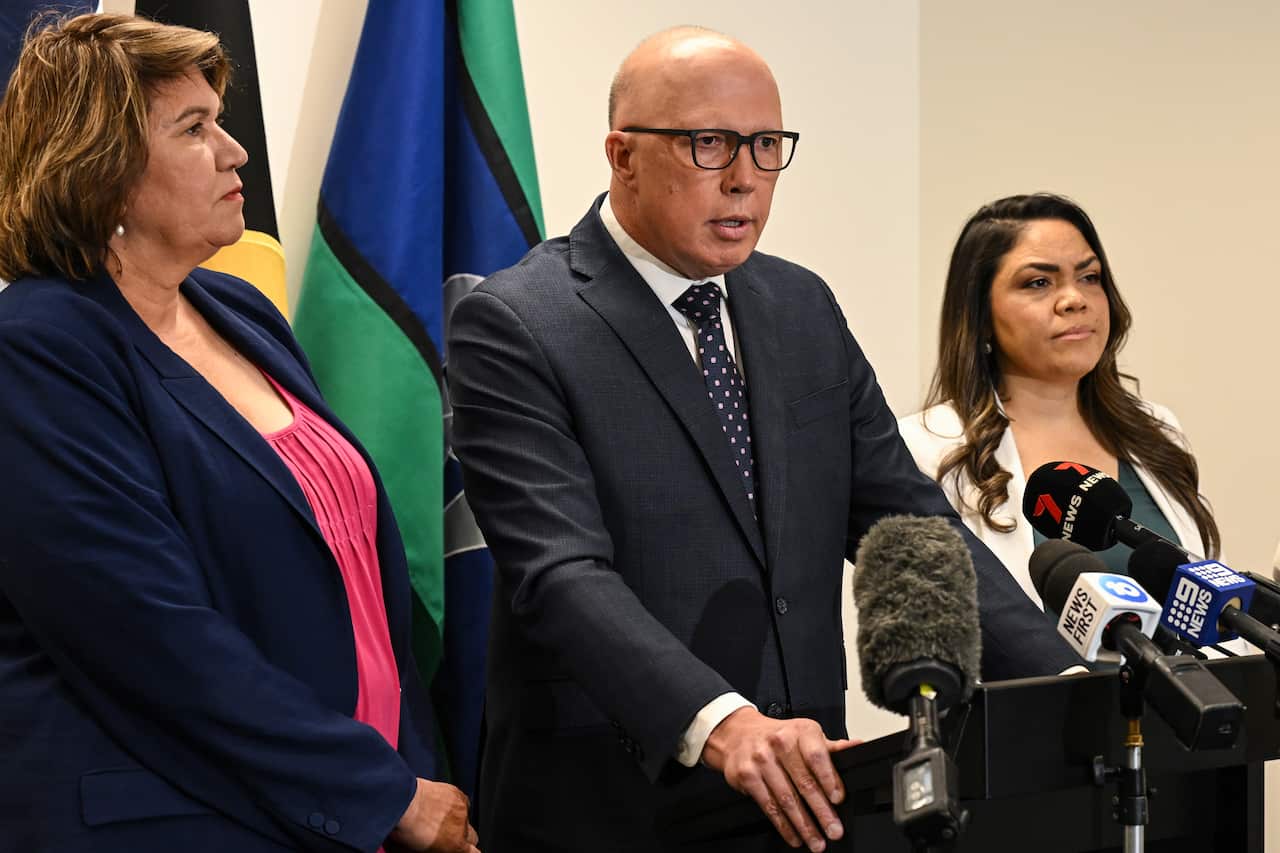 Man and two women stand at podiums in front of Aboriginal and Torres Strait Islander flags.