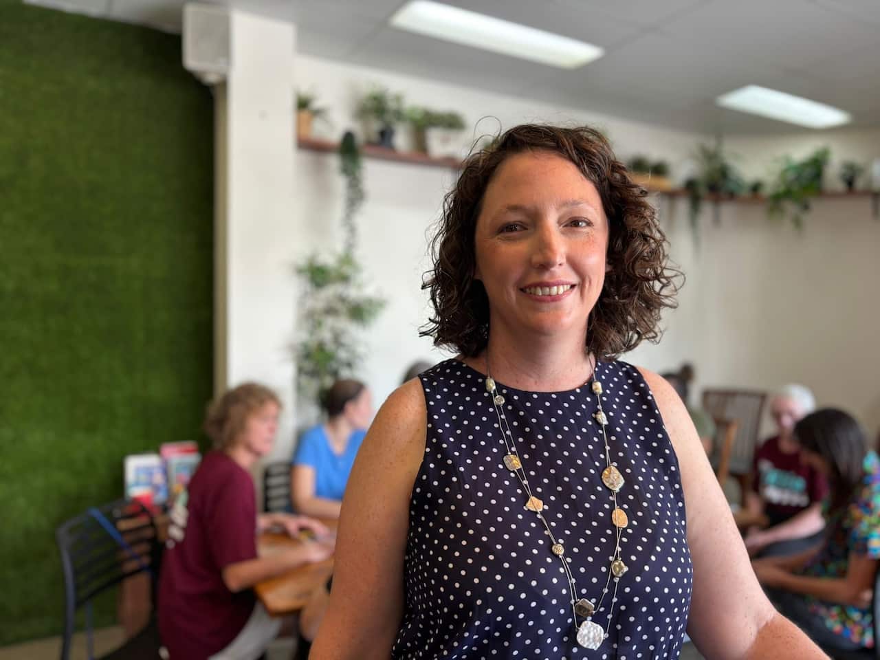 A woman in a polka dot dress, standings smiling in a cafe.