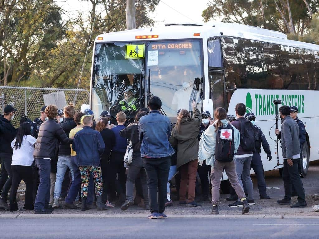 Protesters gather in front of a bus, stopping it from moving forward. 