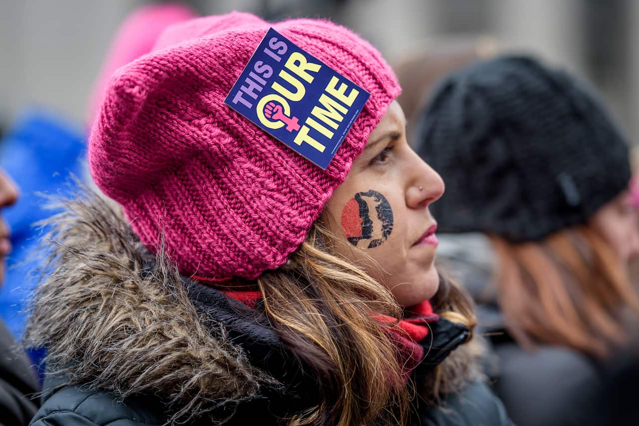 A young woman wearing a pink hat with a sticker attached to it that reads "This is our time" at a protest.