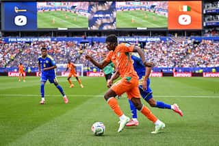 A footballer wearing an orange shirt and shorts dribbles the ball down the field past a defender.