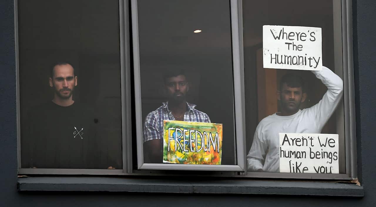 Three men standing at a window with signs including one saying 'Where's the humanity?'