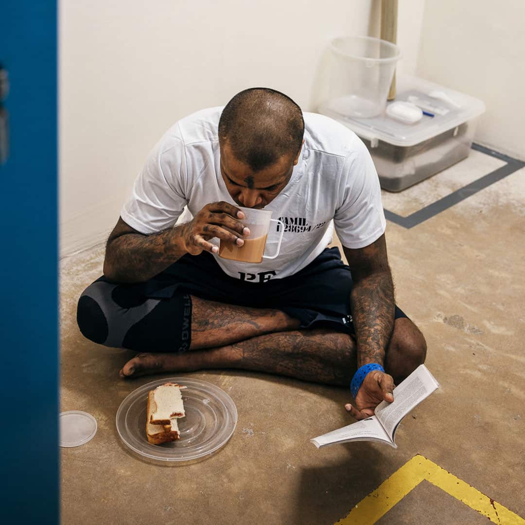 A man sits cross-legged on the floor of a prison cell. In his left hand, he holds an open book, while drinking from a plastic mug in his right hand. There's a plastic plate with slices of bread in front of him