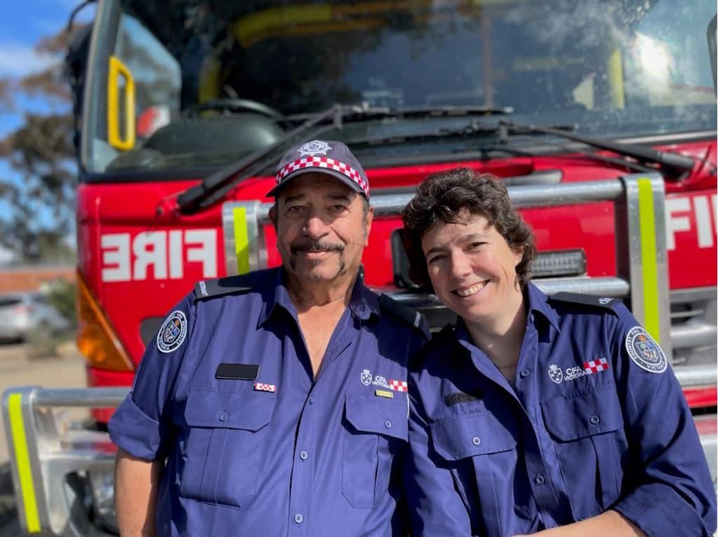 A man and a woman dressed in blue uniforms stand in front of a fire truck