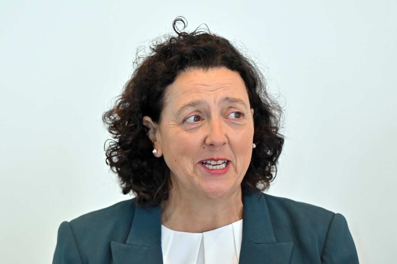 A woman with dark curly hair, wearing a turquoise blazer, is speaking.