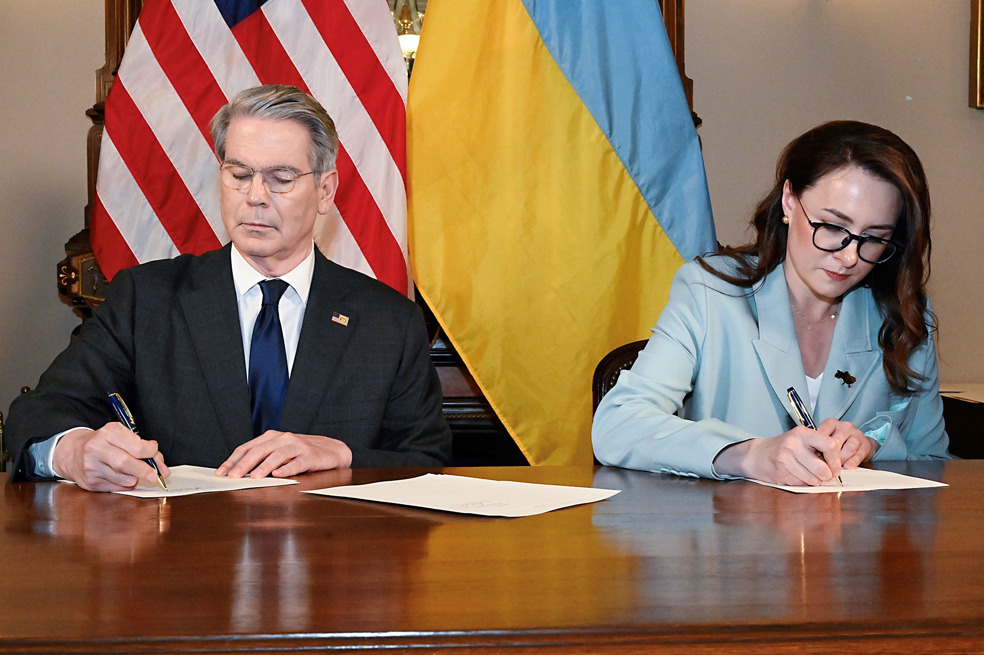 A man and a woman sitting at a desk in front of US and Ukrainian flags, signing documents.