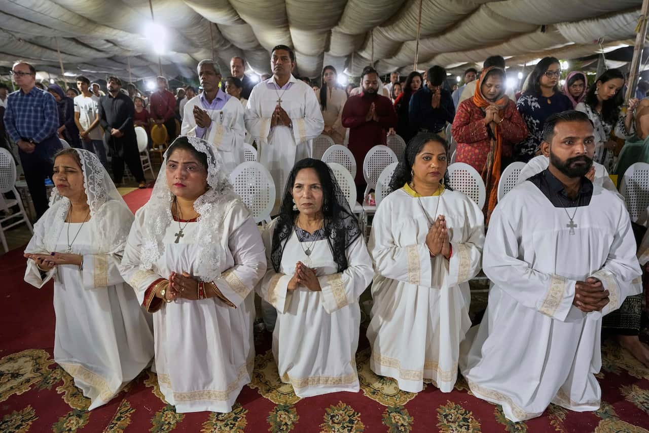 Four women and one man, all wearing long white robes, kneel to pray with more devotees standing behind them.