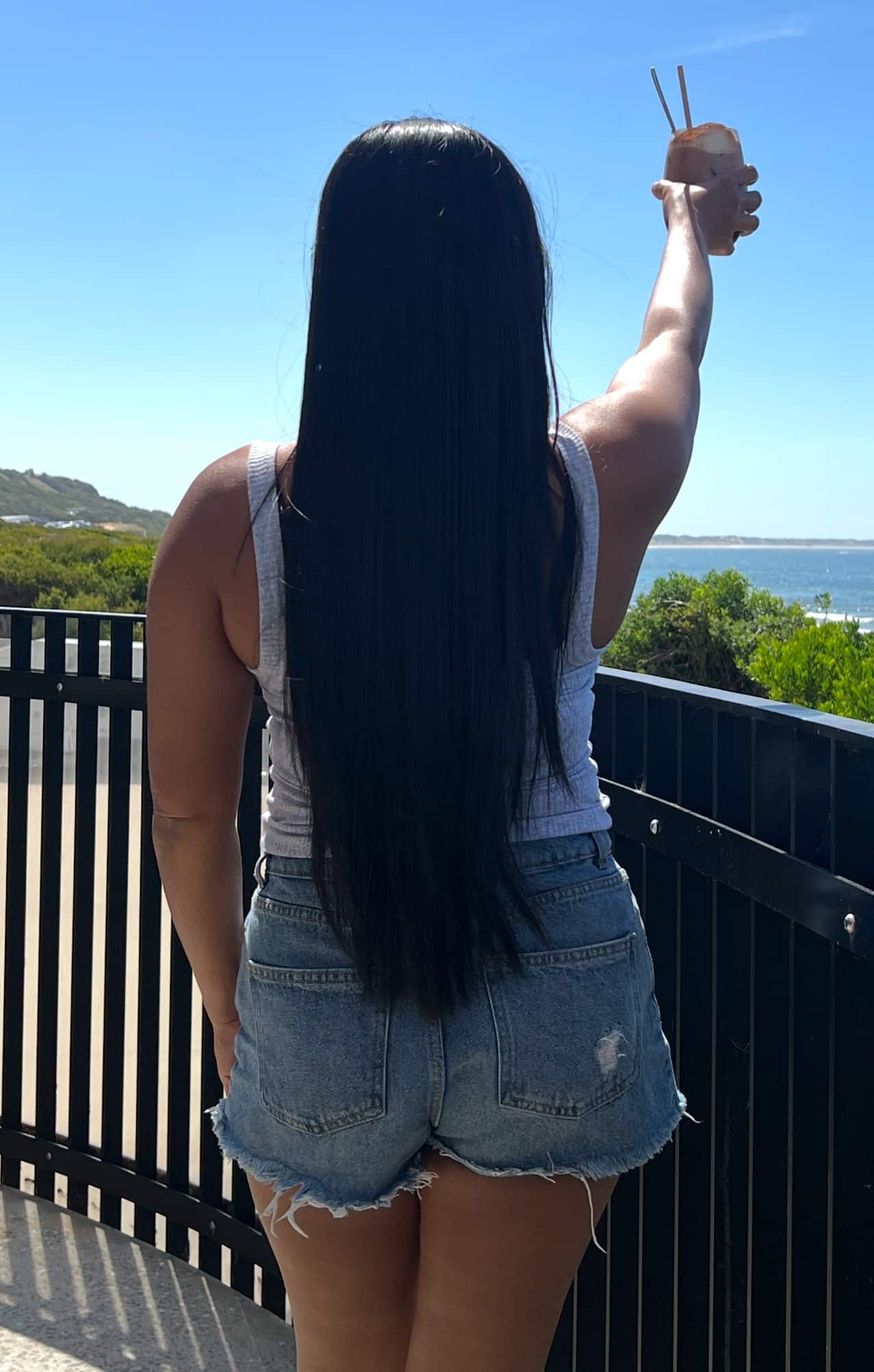 A woman with long black hair, wearing a grey tank top and denim shorts, stands facing a railing overlooking the ocean while raising a coconut drink in her right hand.