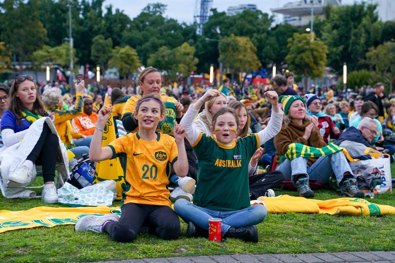 Two children wearing the colours of Australia's football team cheer. A large crowd of people is sat behind them.