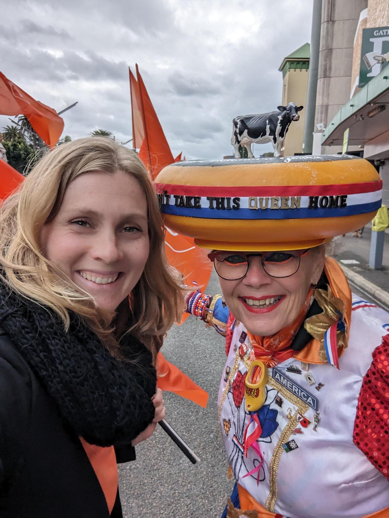 A blonde white woman standing next to another woman wearing a cheese-shaped hat on her head.