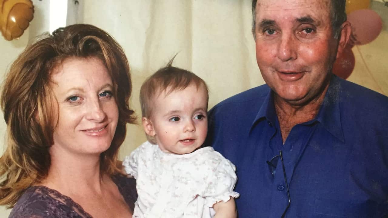 a film photo of a family a young mum holding a baby in a white dress and an older middle aged man hugging them