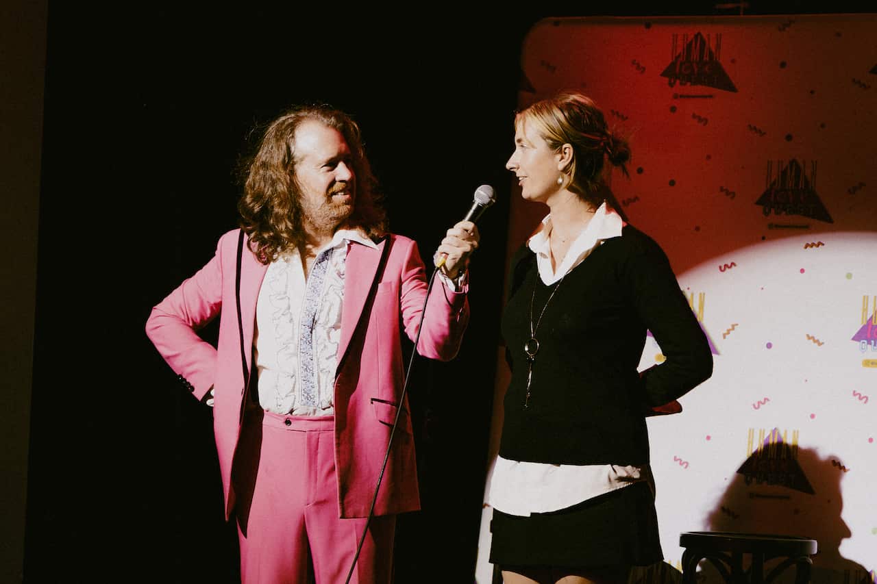 A man wearing a pink suit speaking to a young blonde woman on stage.