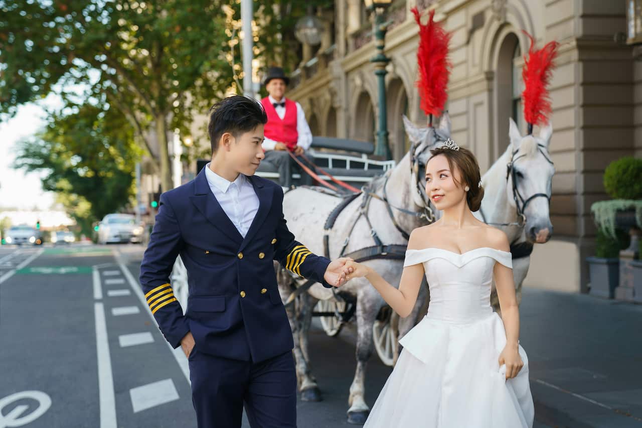 A man in a dark suit and a woman in a white wedding dress walk walk hand in hand through a city street. There is a horse and carriage in the background.
