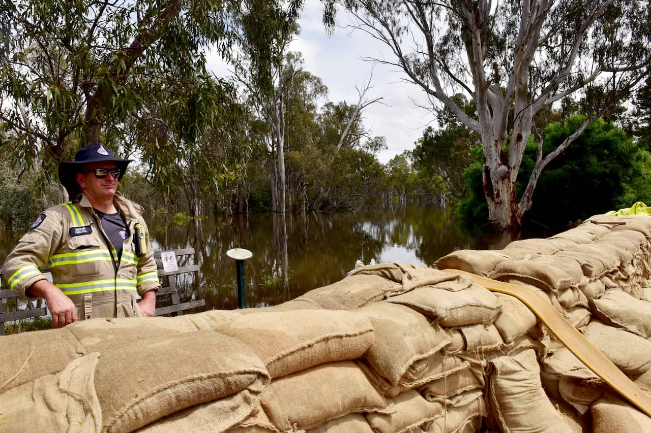 A CFA crew member standing against a fence near a swollen river and flood levee made of sandbags