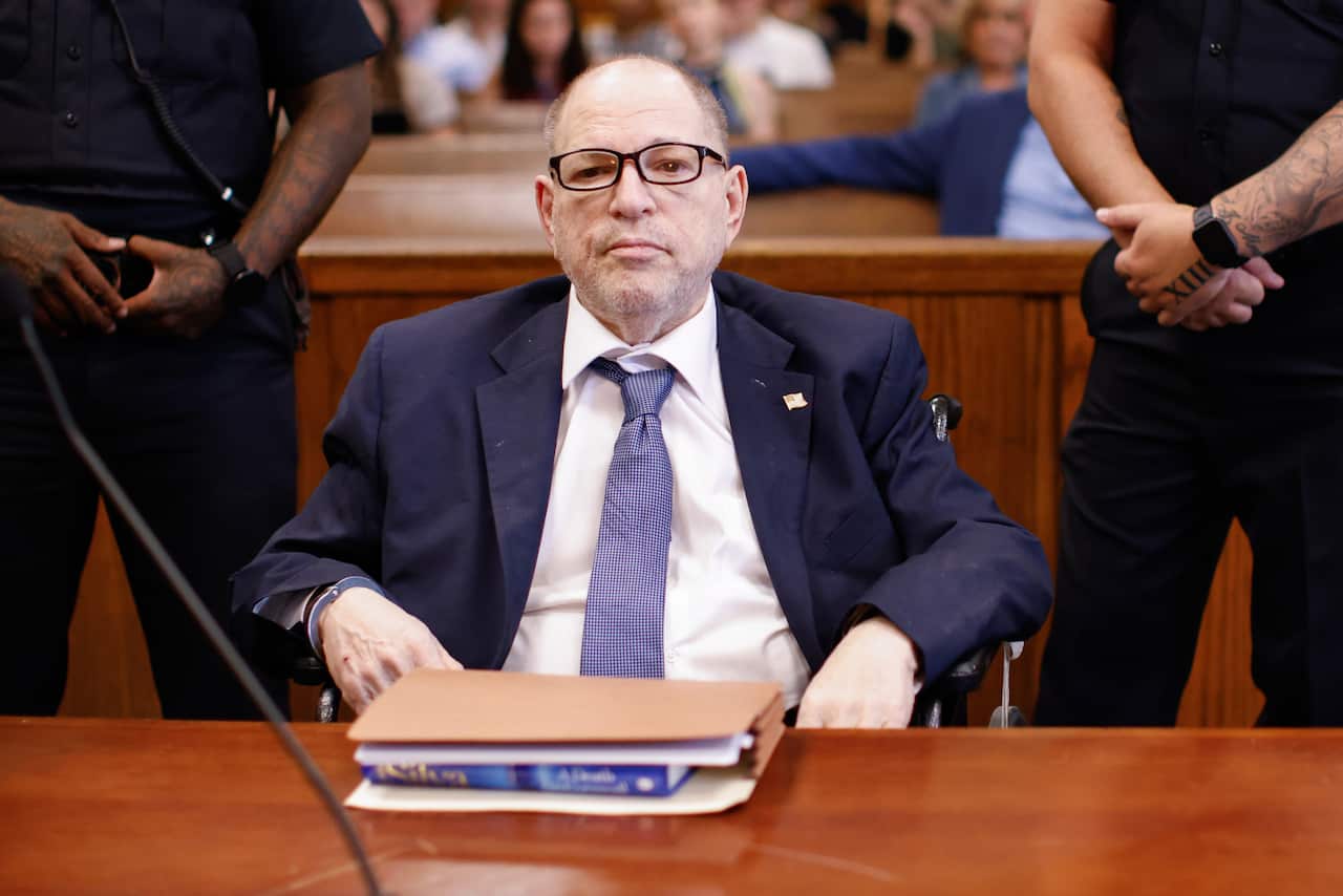 A man in a blue suit is sitting in a courtroom with two guards standing behind him.