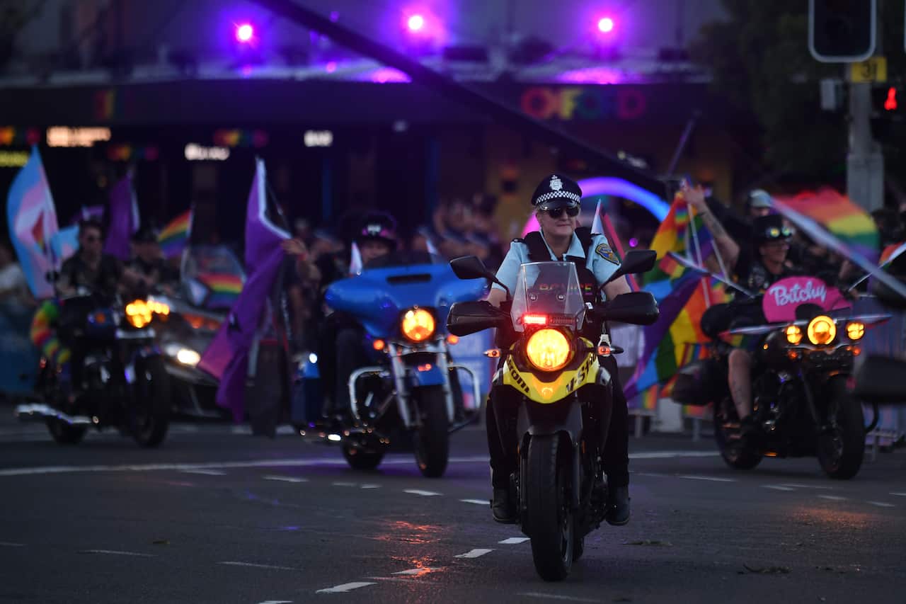 A woman in a police uniform riding a motorbike followed by several others waving rainbow pride and trans flags. 