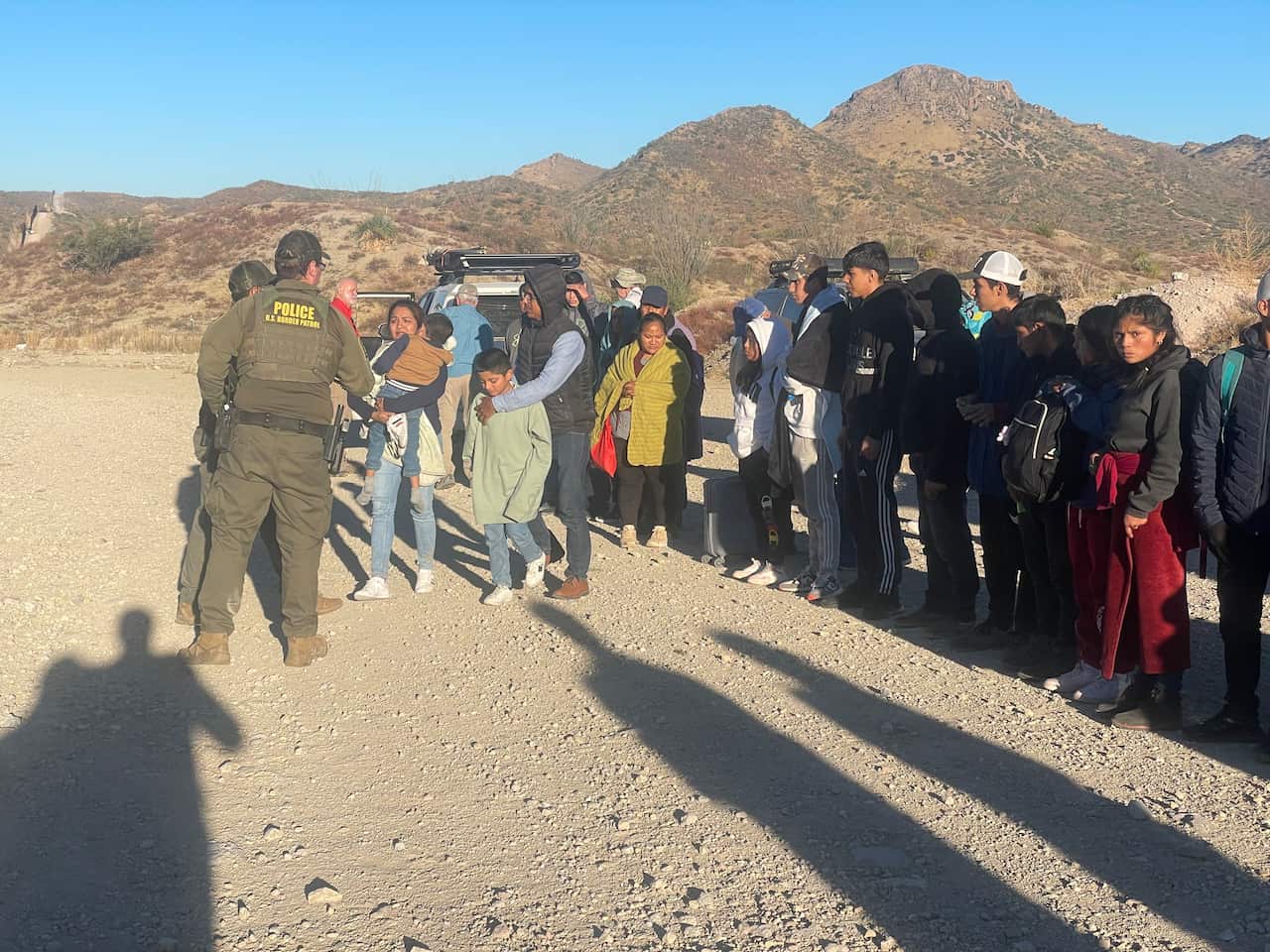 Two border patrol officers speak to migrants, who are standing in a line in the desert.