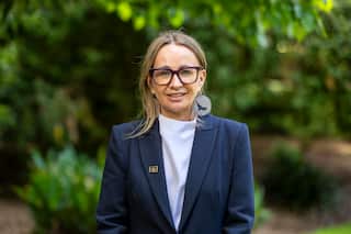 A woman in a navy blue blazer and glasses stands outside.