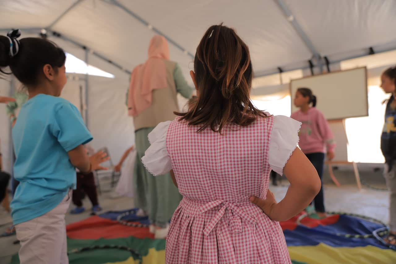 The back of a child wearing a pink gingham dress in a room with other children. 