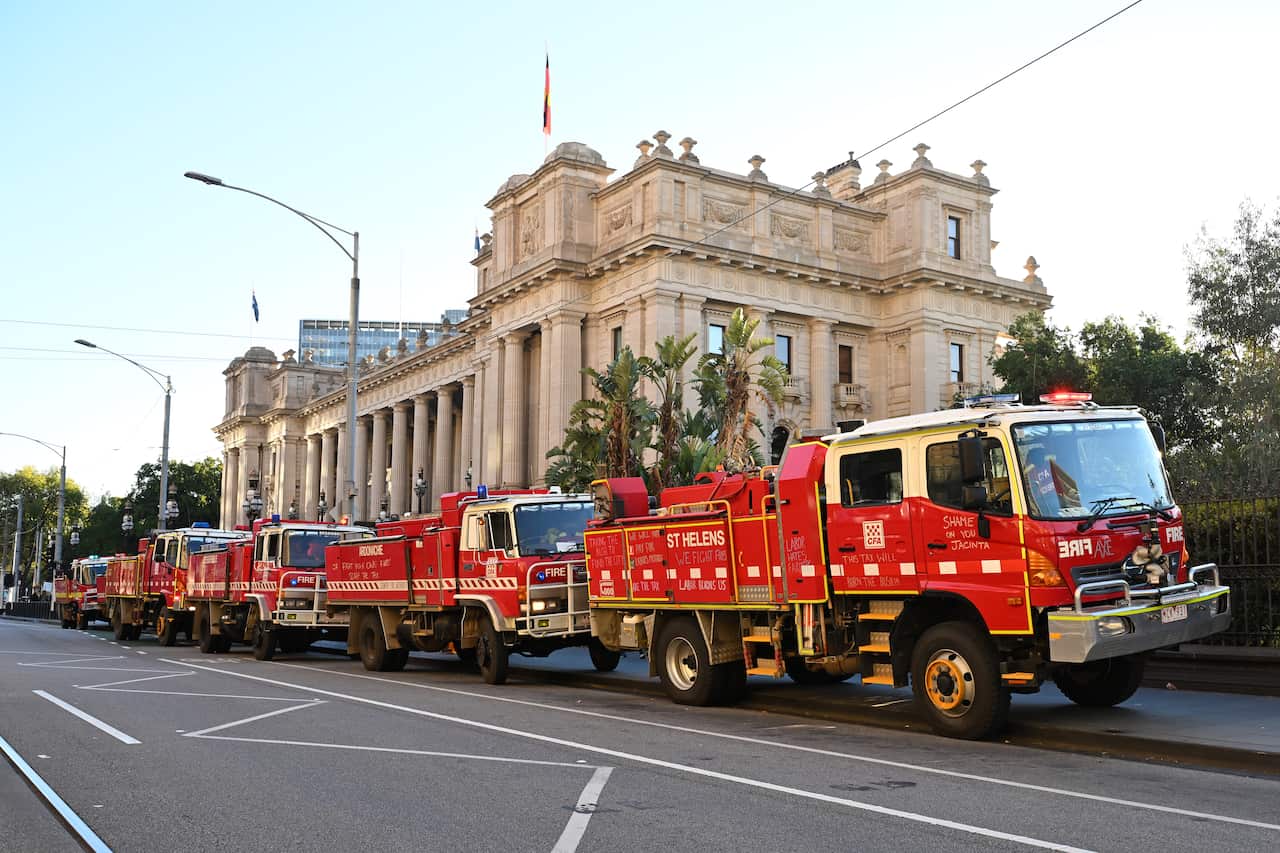 A low-angle, eye-level shot shows a line of red fire trucks parked on a city street. In the background, a large, ornate building with columns and a flag is visible.