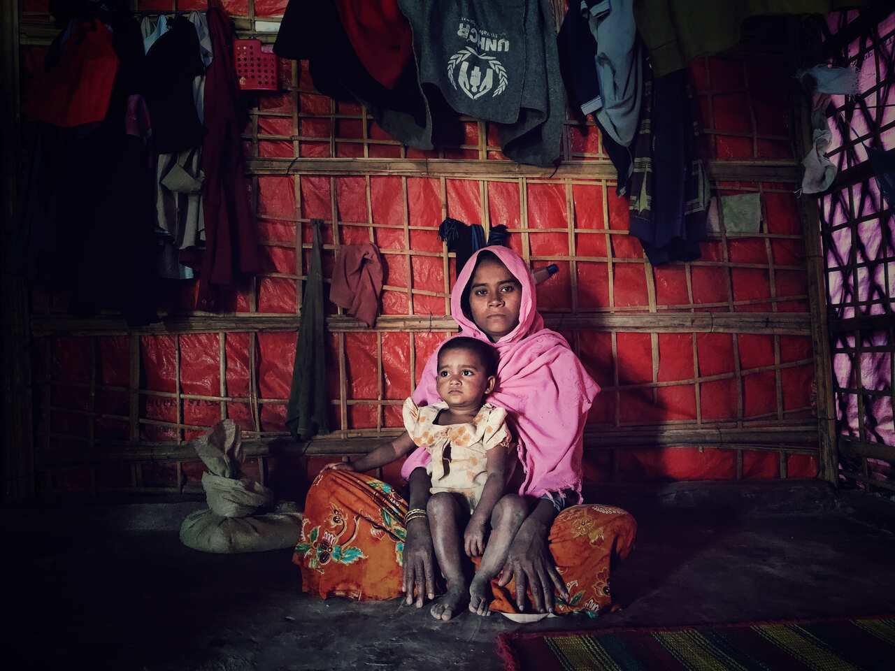 A woman in a pink shawl looking straight at the camera with a child in her lap sitting inside a bamboo-made shelter in a refugee camp.