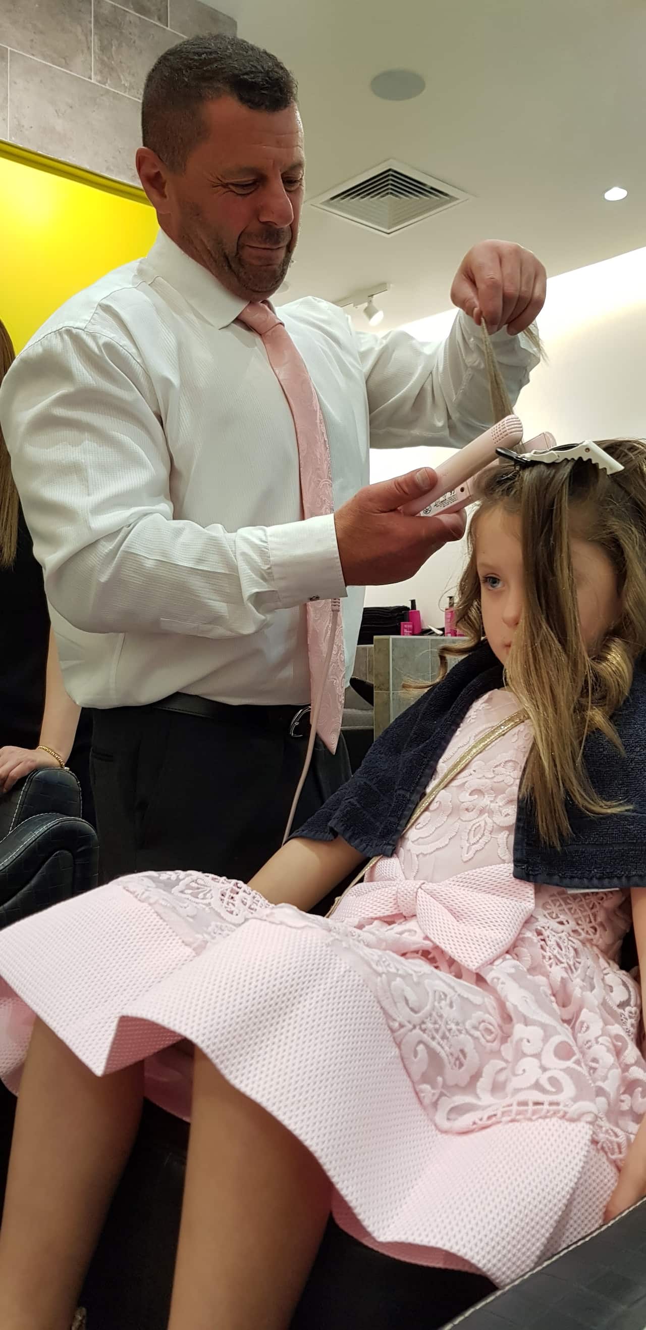 A man stands as he styles the hair of his daughter, who is sitting in a chair.