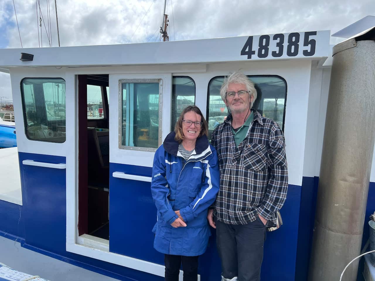 A man and a woman smiling as they stand on a dock in front of a boat.