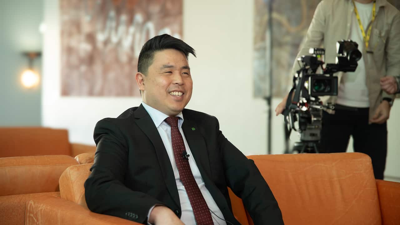 A man in a black suit, sits smiling on an orange couch.