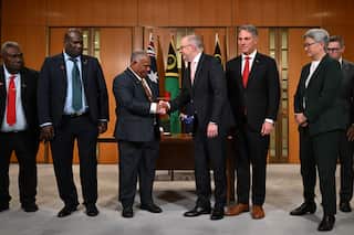 Australian Prime Minister Anthony Albanese shakes hands with Jotham Napat, Prime Minister of Vanuatu, surrounded by officials and the flags of both nations in a wood-paneled room.