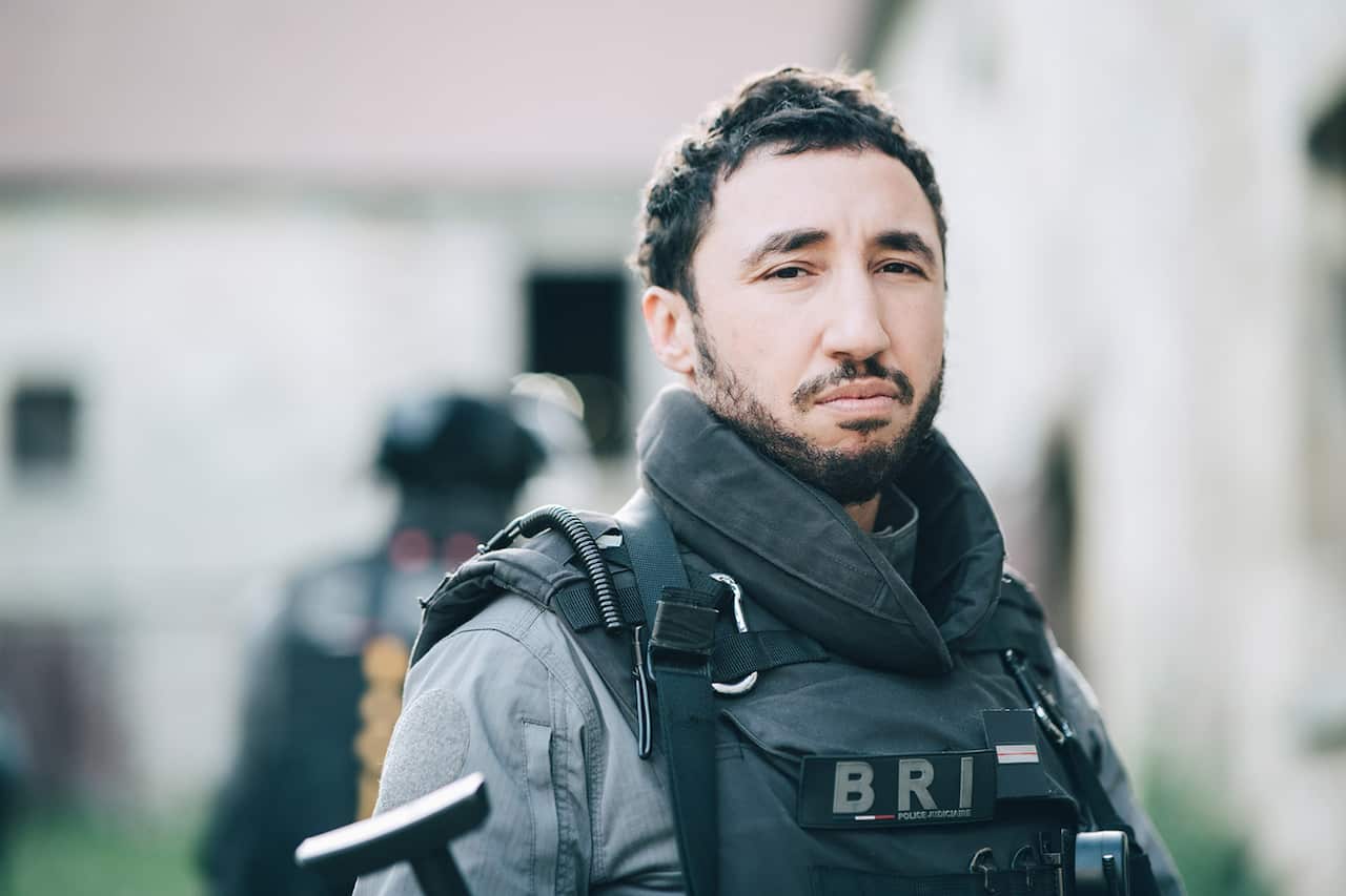 A man in heavy police protective wear looks sideways at the camera.