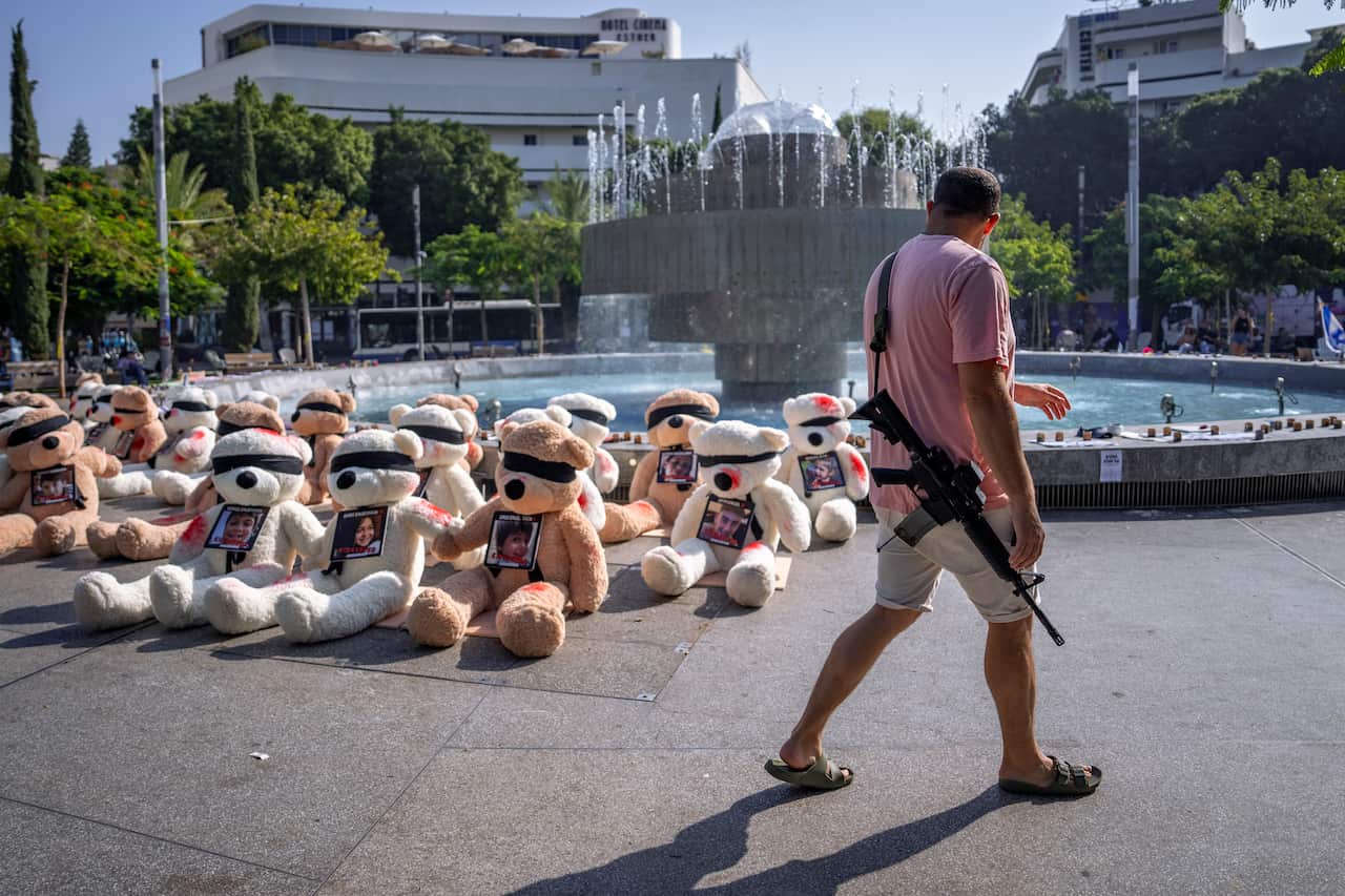 A man with a gun walks past dozens of blindfolded teddy bears.