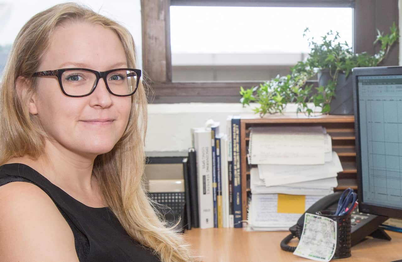 A blonde woman wearing glasses sitting at an office desk and smiling.