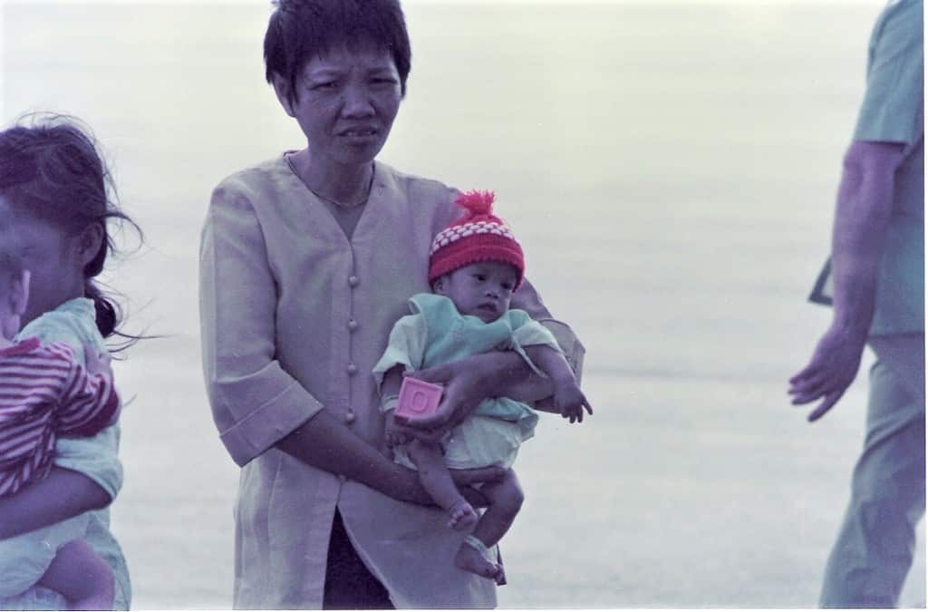 A Vietnamese woman holding a very small baby in a red beanie and green jumper, waiting on an airport tarmac.
