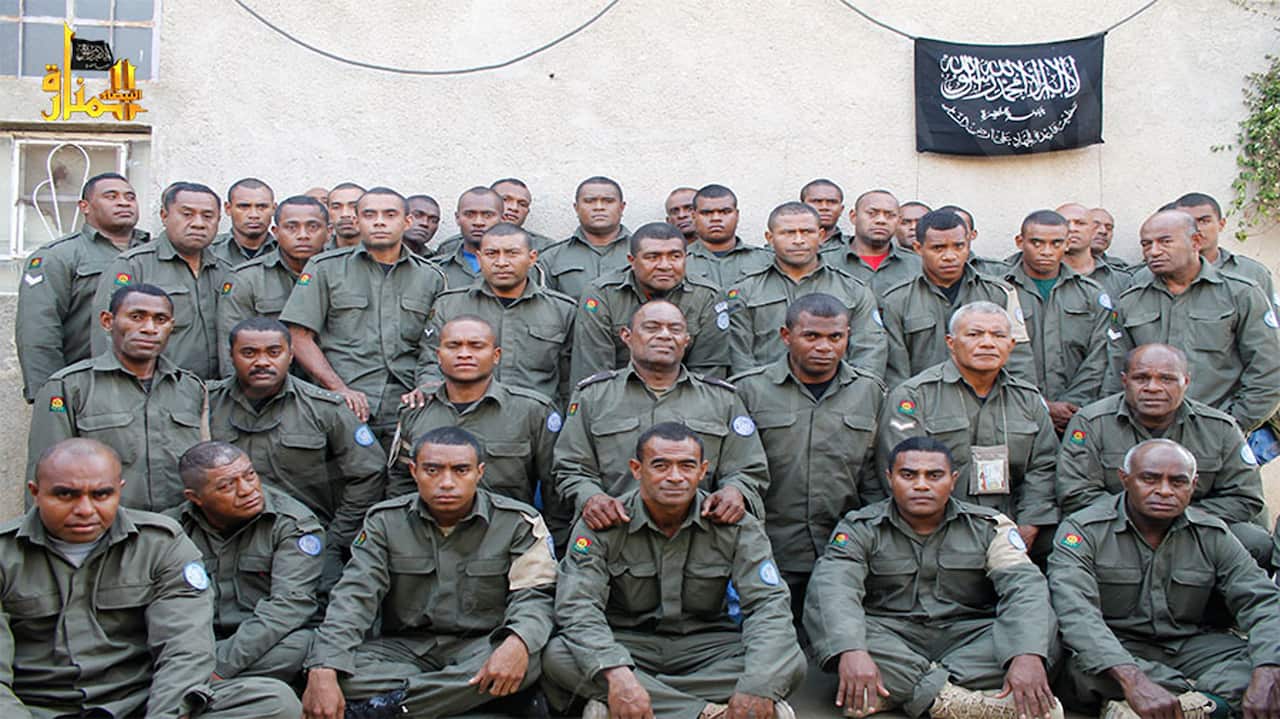 A group of soldiers in olive green uniforms posing for a group photo in front of a plain wall with a black flag hanging in the background.