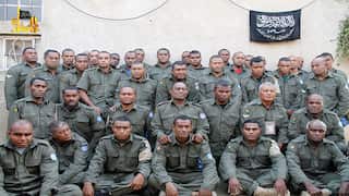 A group of soldiers in olive green uniforms posing for a group photo in front of a plain wall with a black flag hanging in the background.