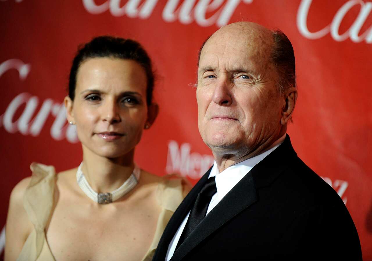 A woman wearing a beige dress and a man wearing a suit standing in front of a red wall.