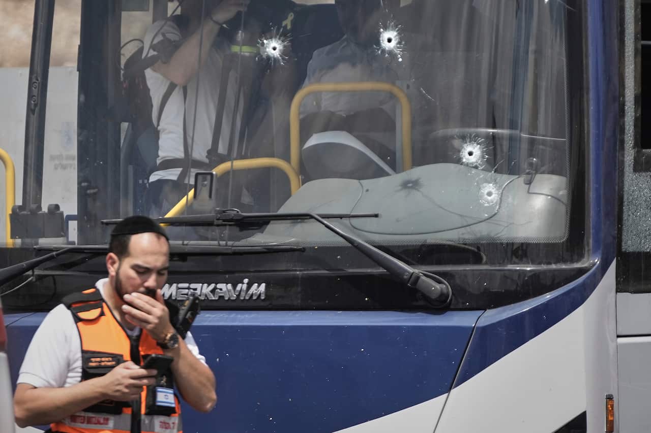 A bus front window with bullet holes in it. A security personnel in front is holding his mouth.