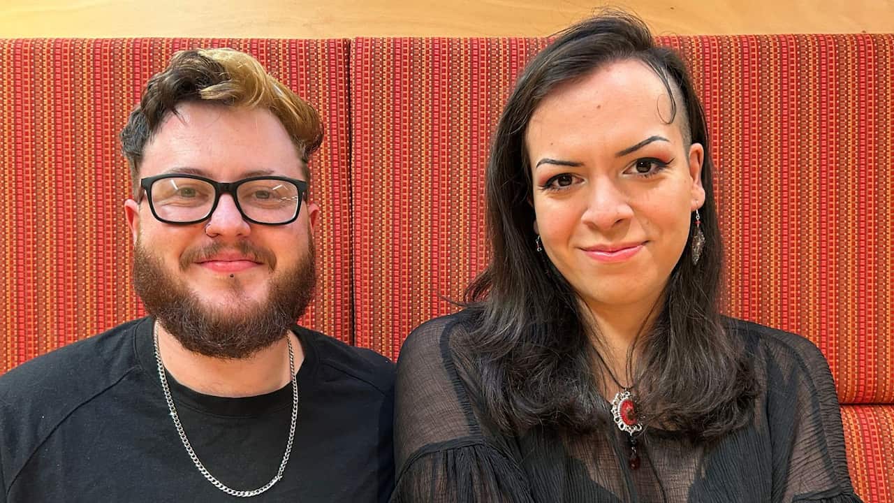 a young man with a beard and glasses sits smiling next to a woman with winged eyeliner and dressed in sheer black in front of an orange background