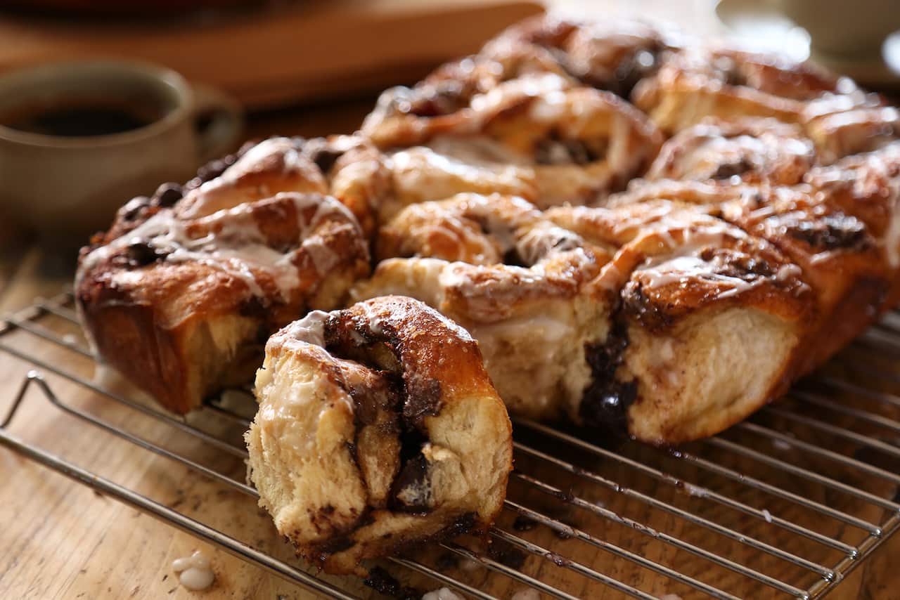 A rectangle-ish set of cooked sticky buns sits on a wire cooking rack, one is torn off the front and sites separately, showing the slighlty melted choc interior and sticky glaze.