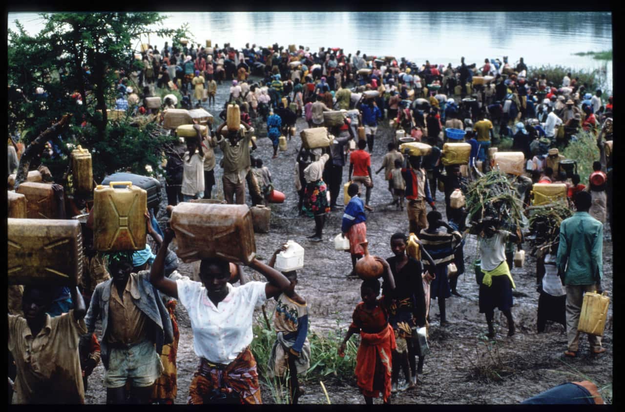 Hundreds of people carrying luggage walk along a river bank