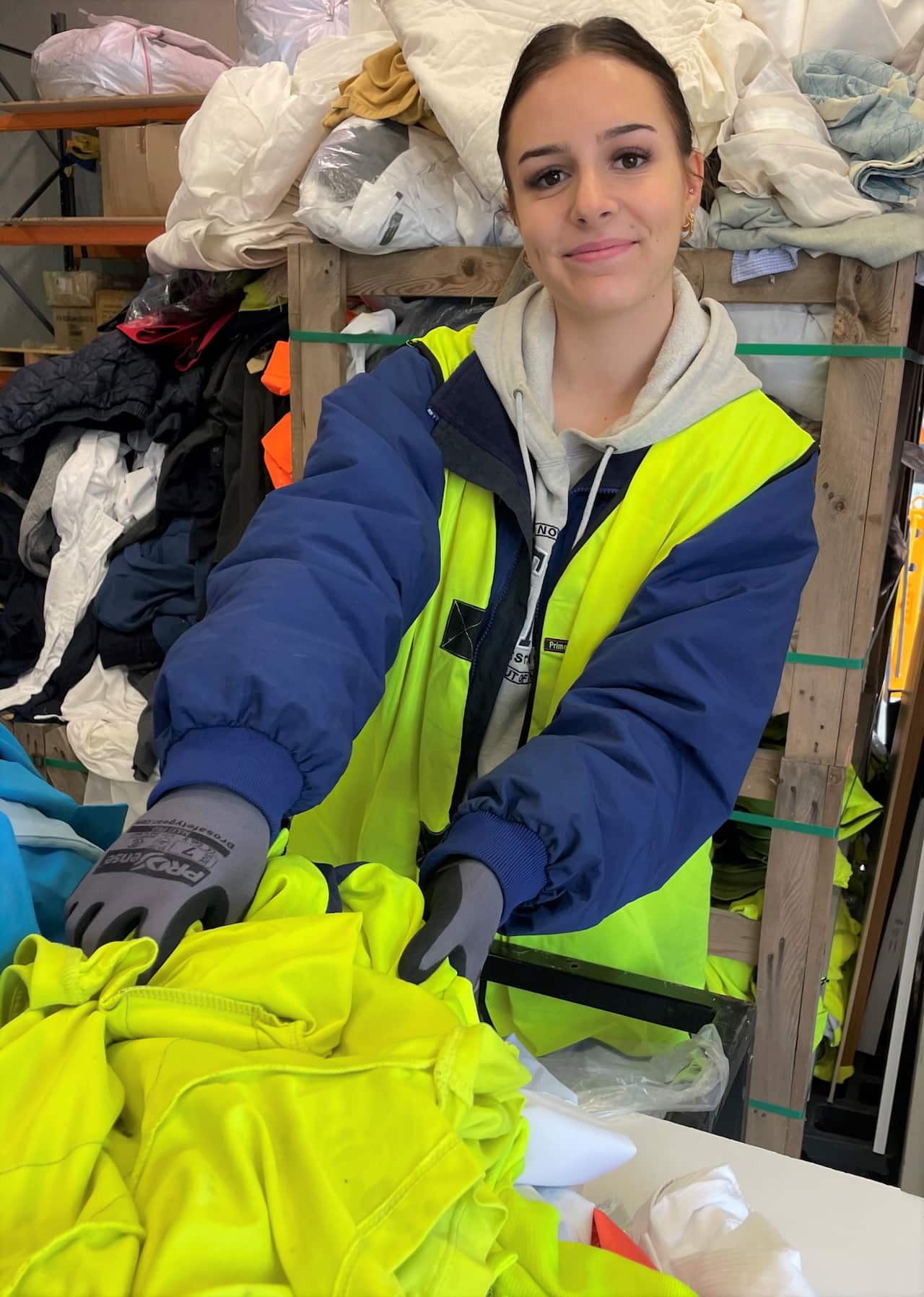 A young woman in a hi-viz vest sorts discarded uniforms at a warehouse. 
