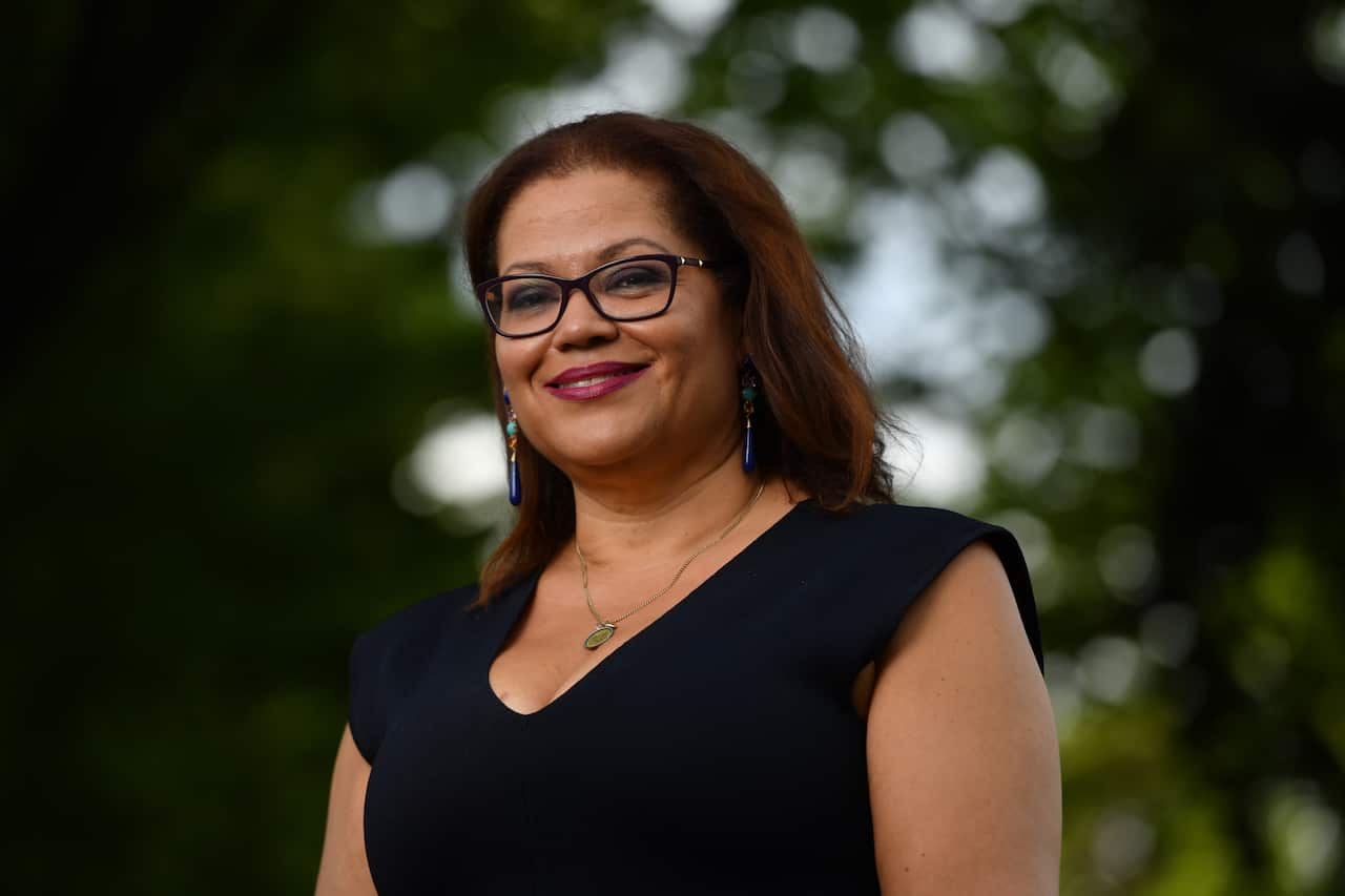 A woman wearing a black dress and glasses looks forward smiling
