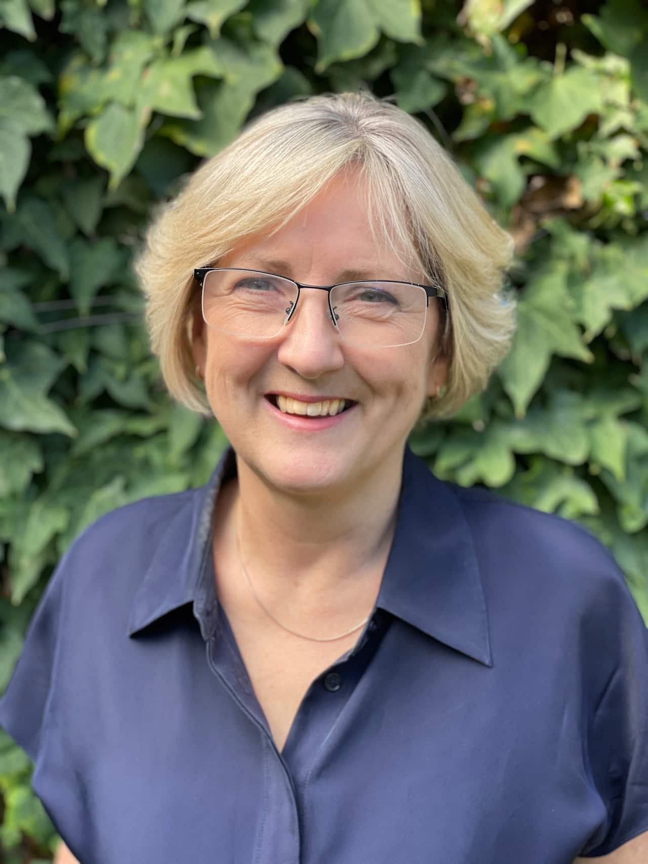 a woman wearing a blue collared shirt, short blonde hair and glasses is smiling 