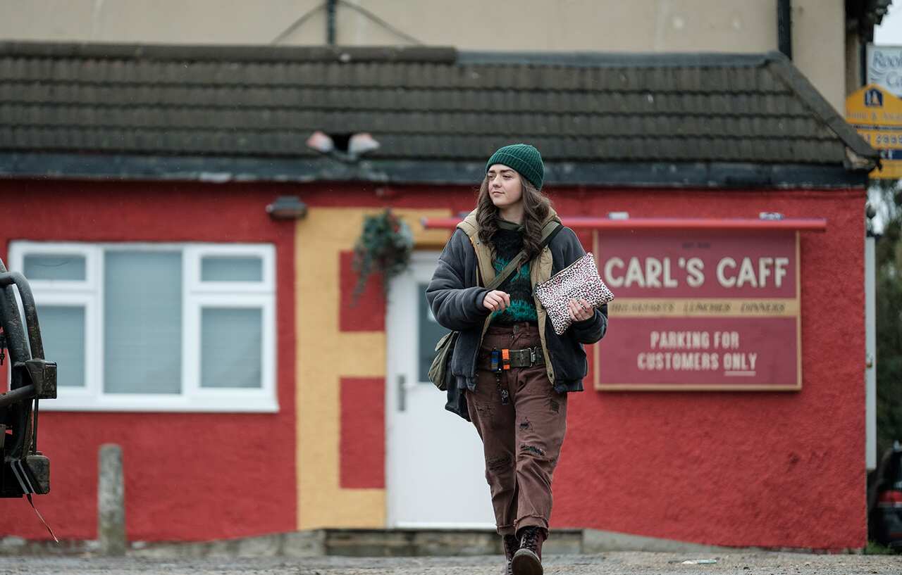 A woman in a dark jacket brown pants, boots and a beanie, with a bag slung across her body, walks away fron a building with a sign on it that reals 'Carl's Caff'.