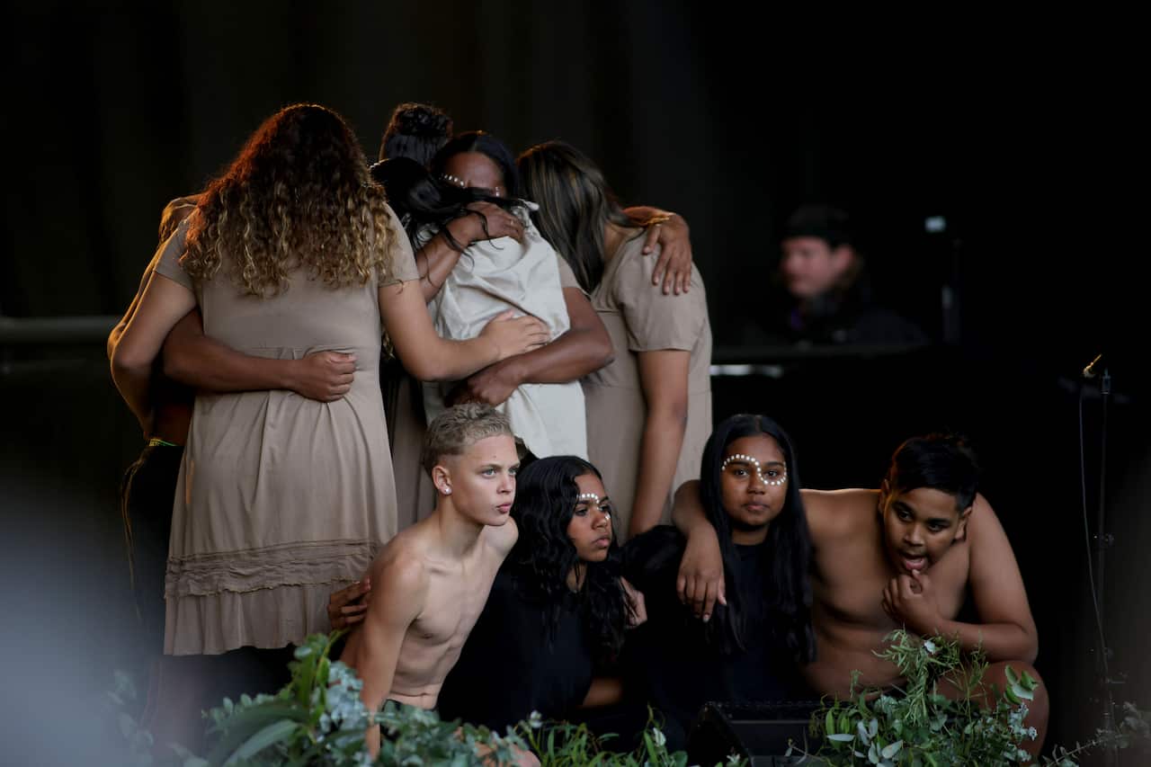 children in traditional paint crouch together on stage, while a group of women standing behind embrace each other. 